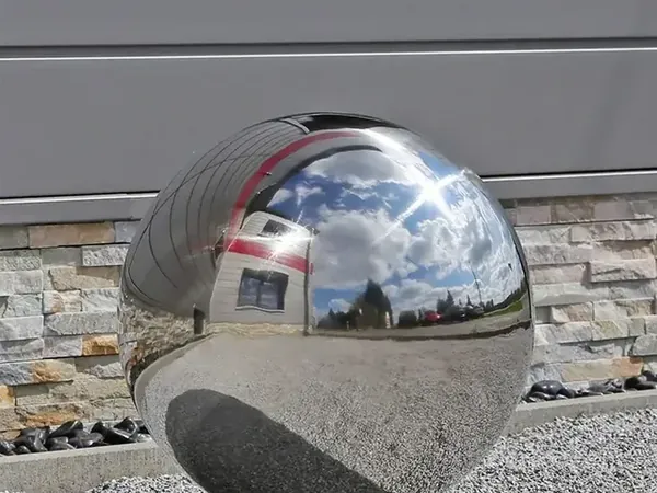 Polished stainless steel sphere on gravel reflecting sky, clouds, and nearby buildings against a stone-clad wall — Ball-Sculpture (China)