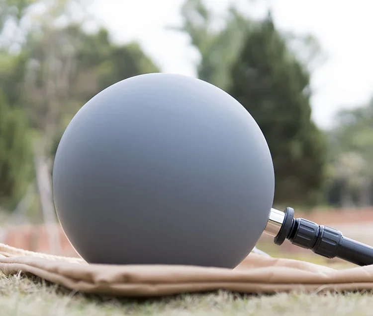 Gray spherical product with black hose fitting resting on a tan cloth on grass, blurred trees behind