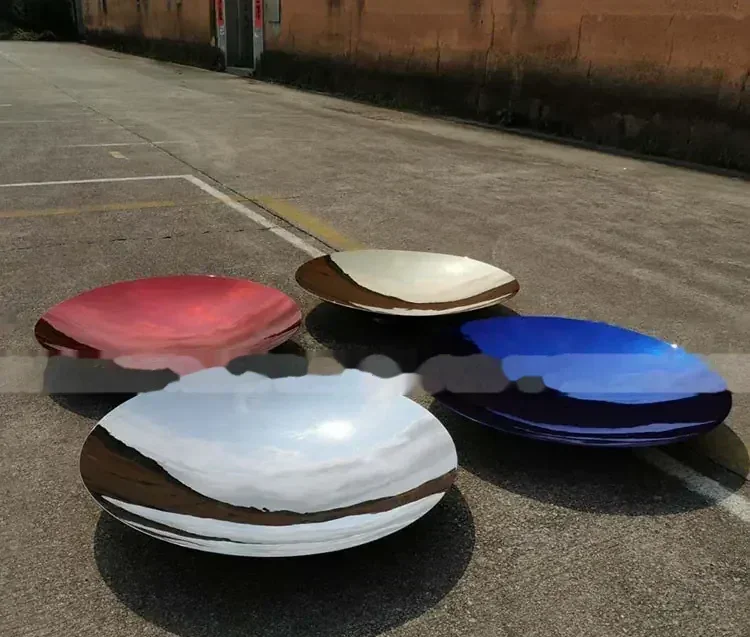 Four polished concave metal dishes in red, gold, blue and mirror finishes placed on outdoor pavement