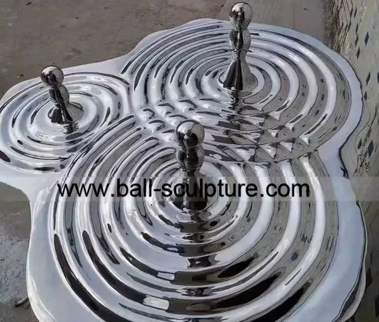 Mirror-polished stainless steel water-ripple sculpture with three sphere-topped finials on a concrete floor