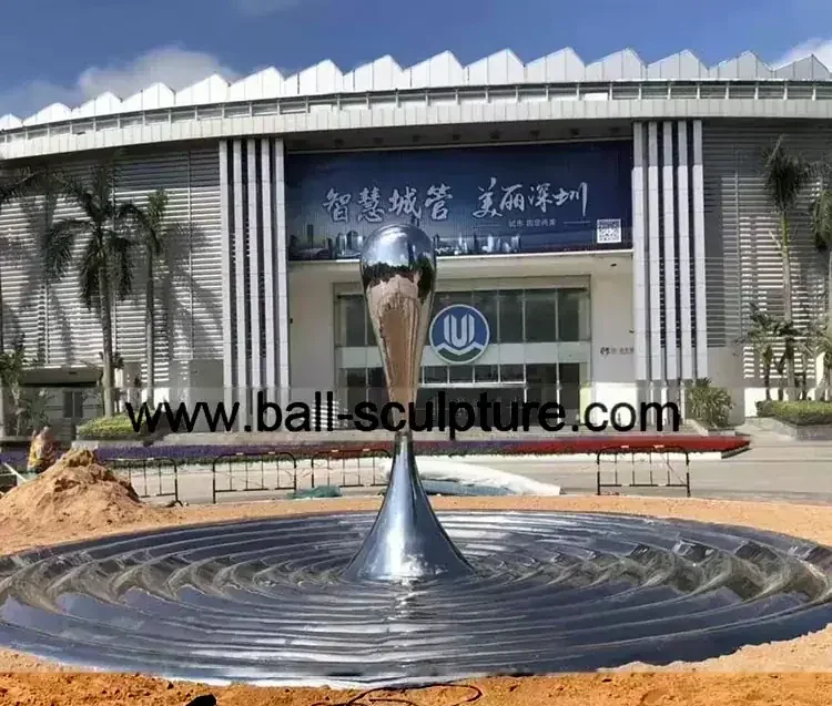 Large, polished metal sculpture resembling a water droplet splash, situated outdoors in front of a modern building.