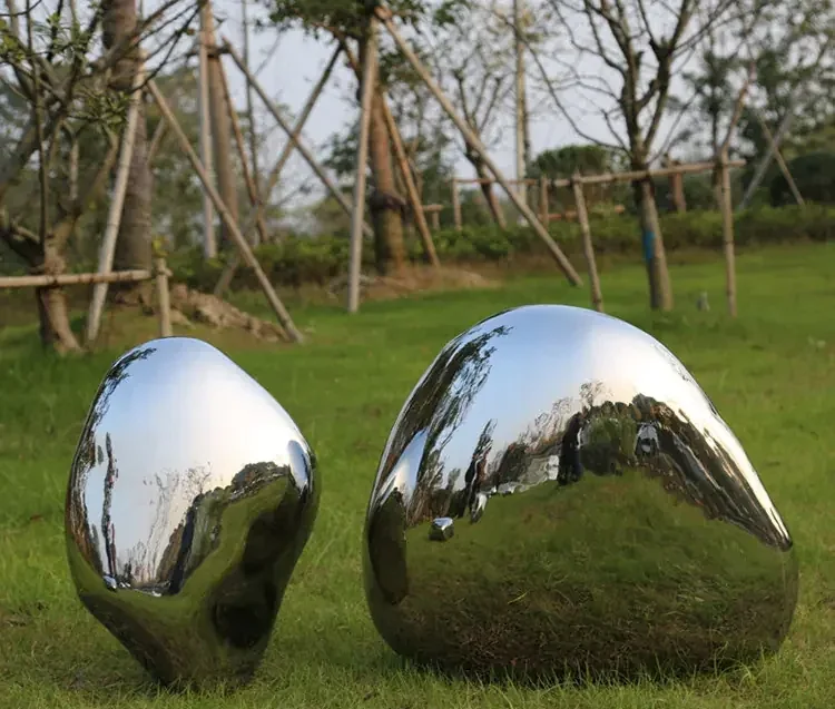 Two polished mirror-finish stainless steel pebble sculptures displayed on a grass lawn with trees behind