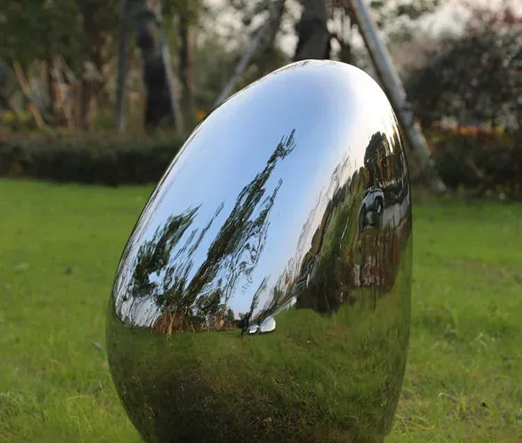 Polished stainless steel egg-shaped sculpture with mirror finish displayed on a grass lawn