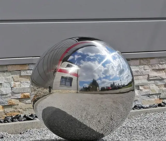 Polished stainless steel sphere on gravel reflecting sky, clouds, and nearby buildings against a stone-clad wall