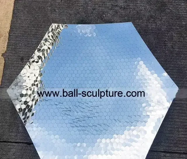 Hexagonal panel covered in small mirror tiles reflecting sky, lying on dark pavement with website URL overlaid