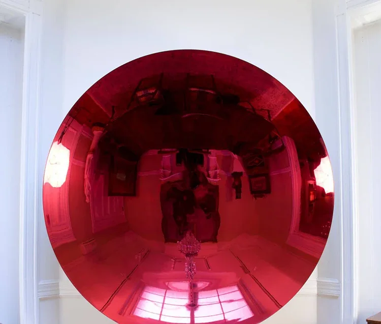 A large red circular mirror mounted on a white wall, reflecting a person taking the photo and the room's interior.