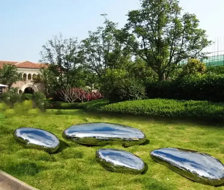 Four polished stainless steel stone-shaped sculptures placed on a green lawn in a landscaped garden.