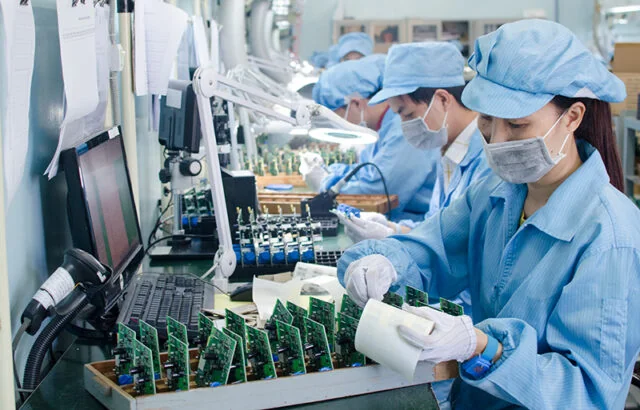Workers in blue uniforms and masks assemble green circuit boards on a factory production line.